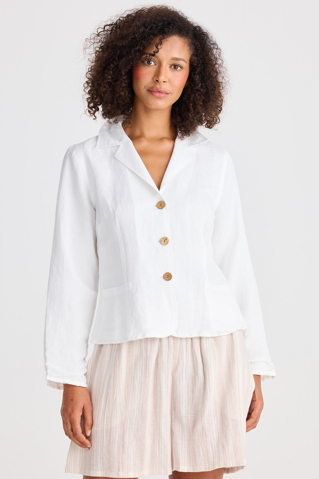 Woman wearing a white jacket with wooden buttons and beige shorts on a white background