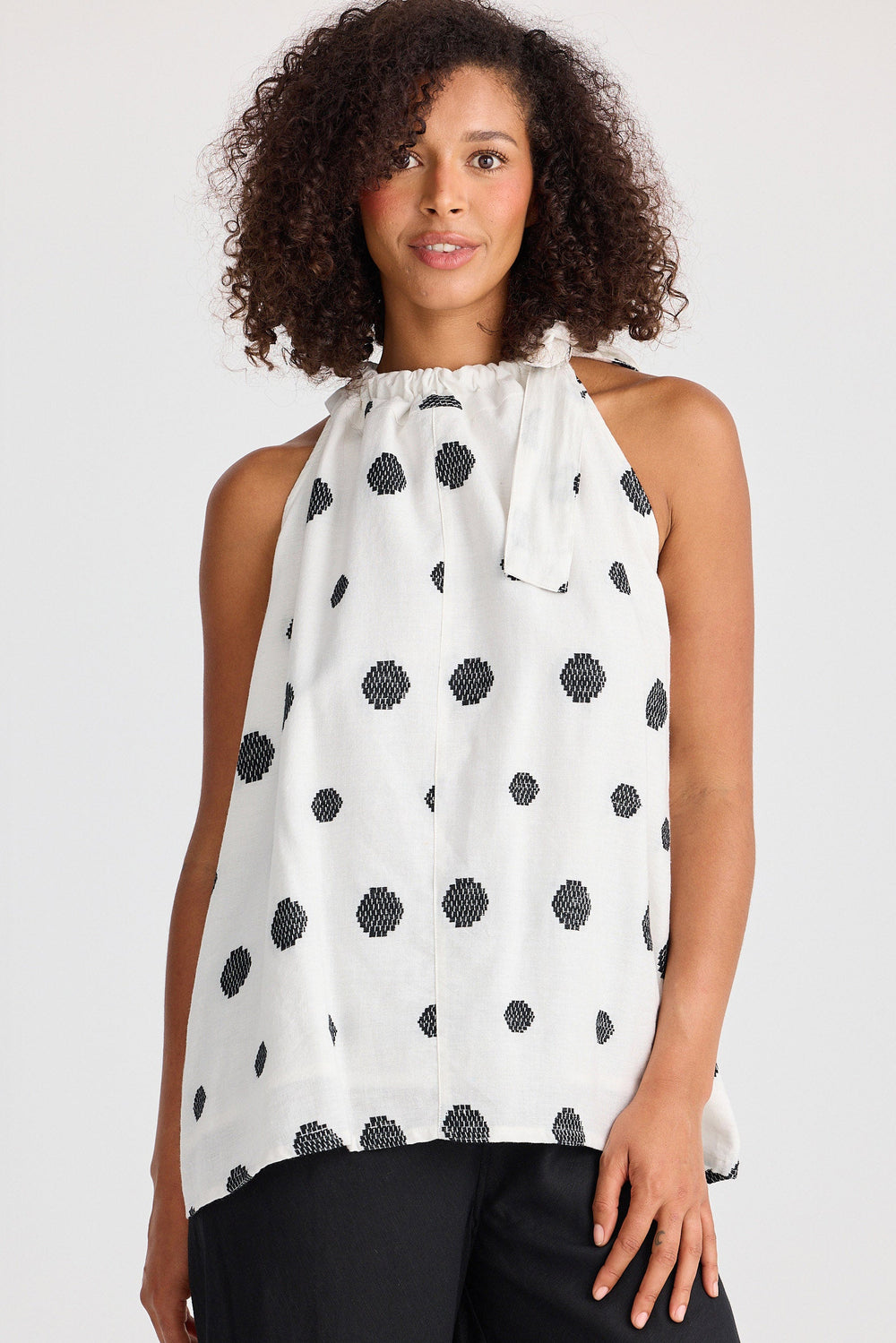 Woman wearing a white sleeveless top with black polka dots and black pants on a white background