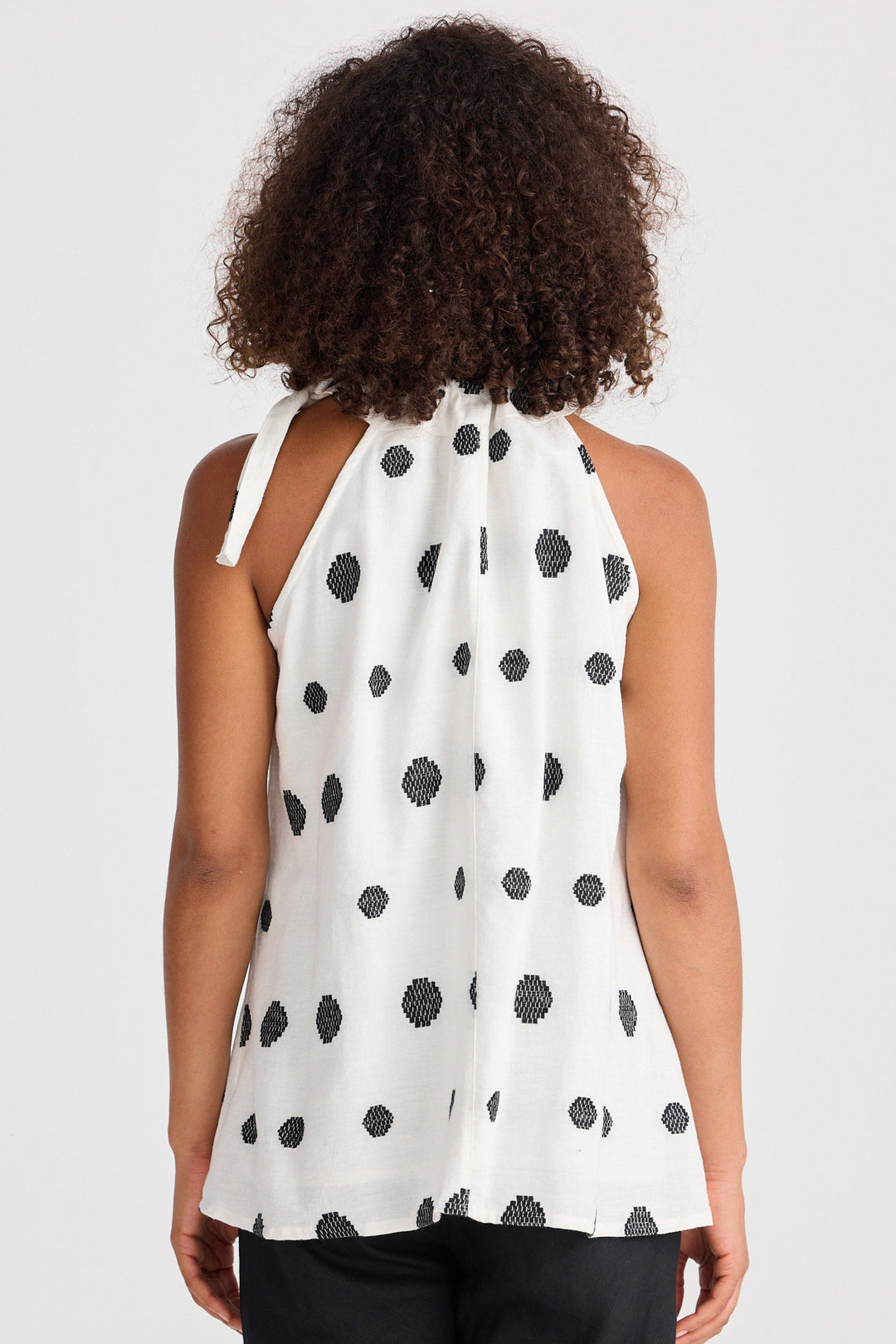 Woman wearing a white sleeveless top with black polka dots and black pants on a white background