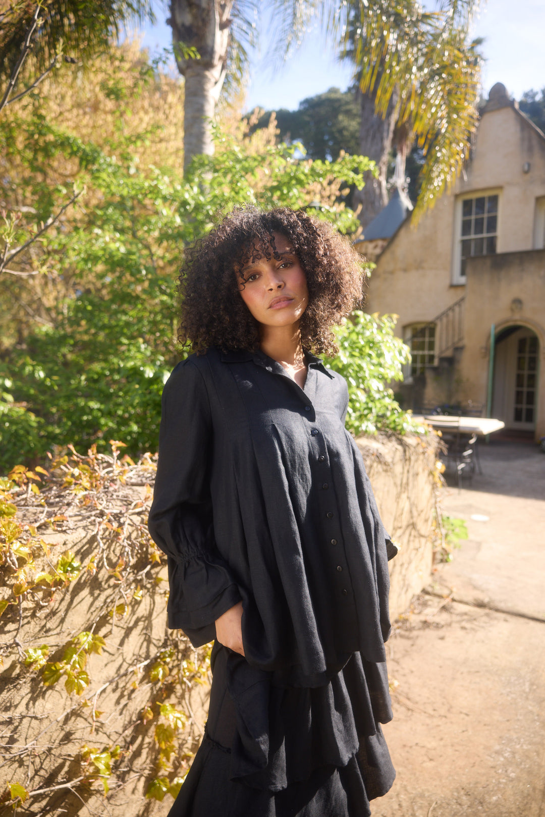Woman in a black outfit standing outdoors with trees and a building in the background