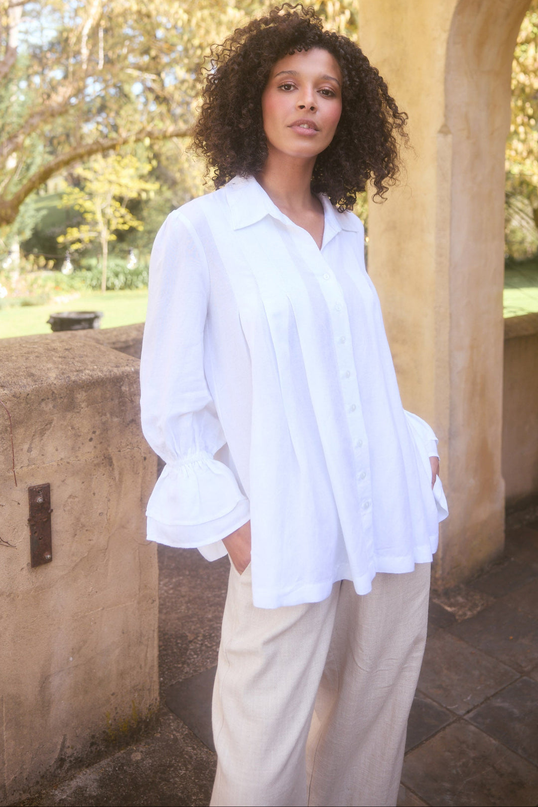 Woman in a white shirt standing in a garden with trees and stone archway in the background