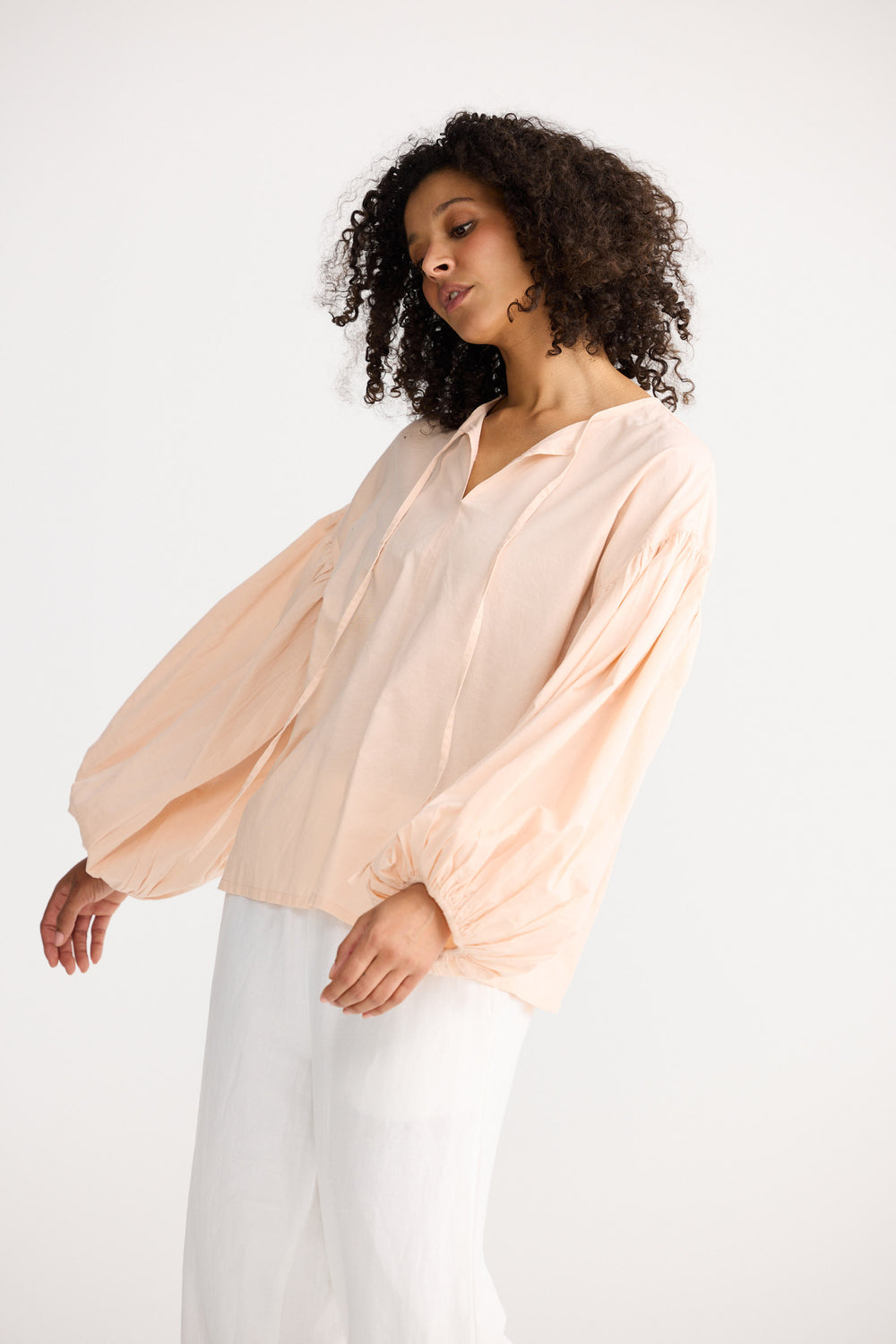 Woman wearing a light peach blouse with long sleeves and white pants on a plain background