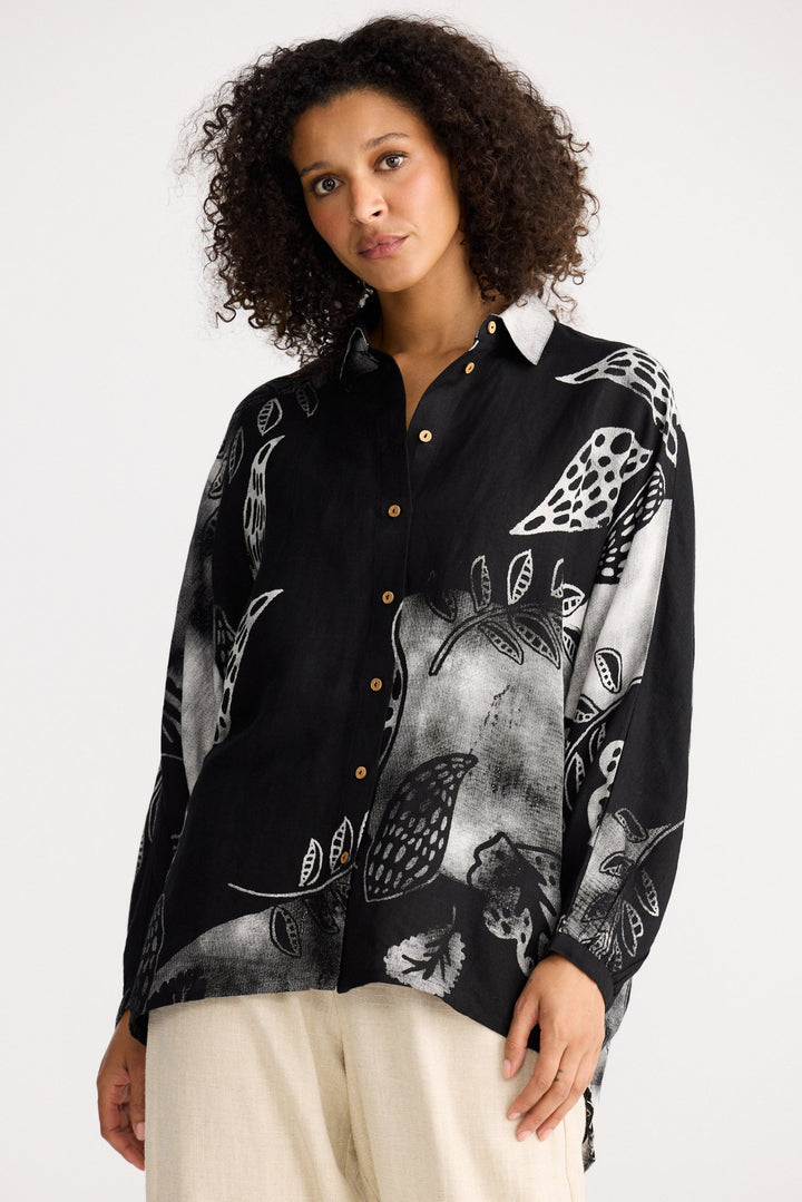 Woman wearing a black patterned shirt with white pants on a plain background