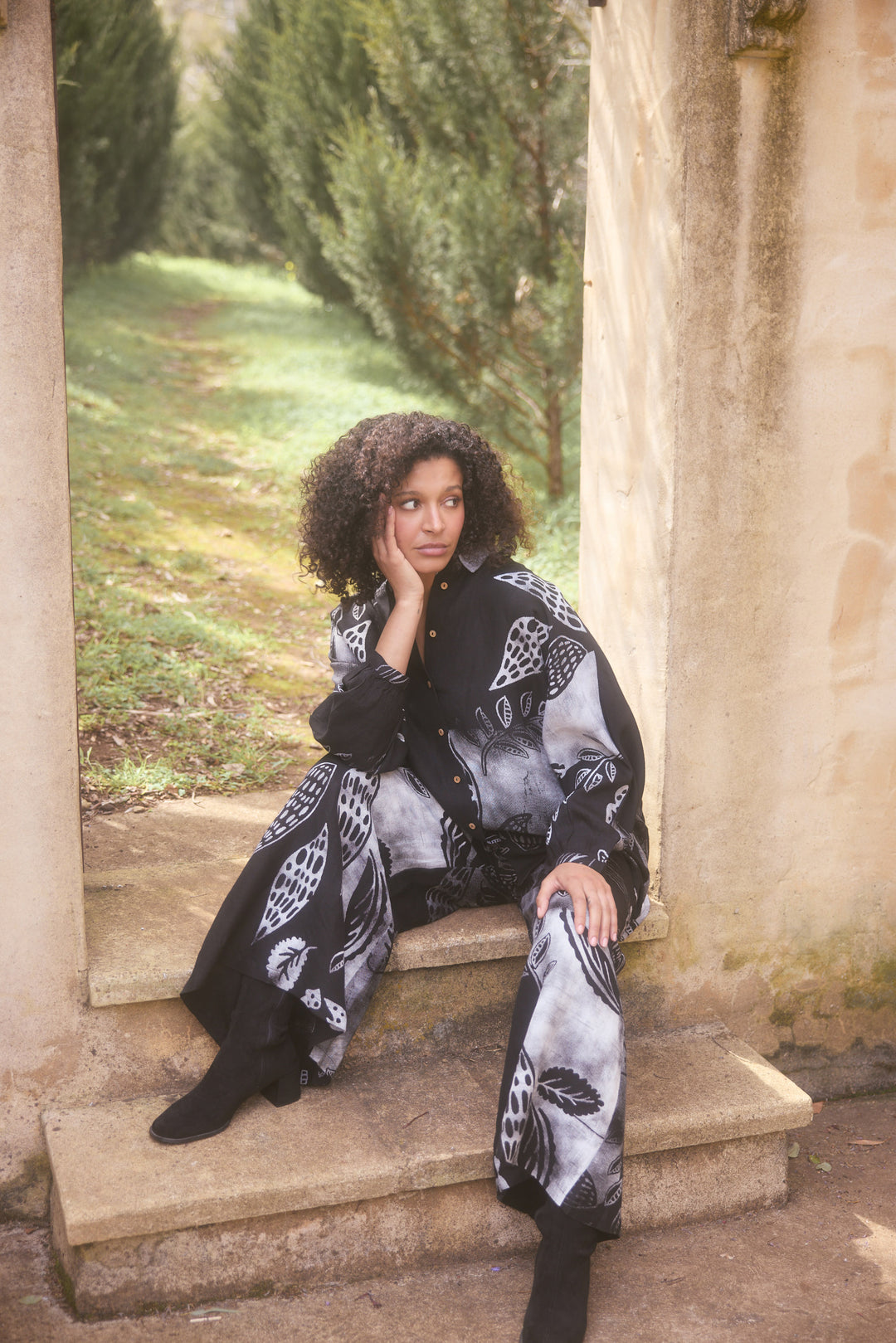 Woman sitting on stone steps in a garden wearing a patterned outfit.