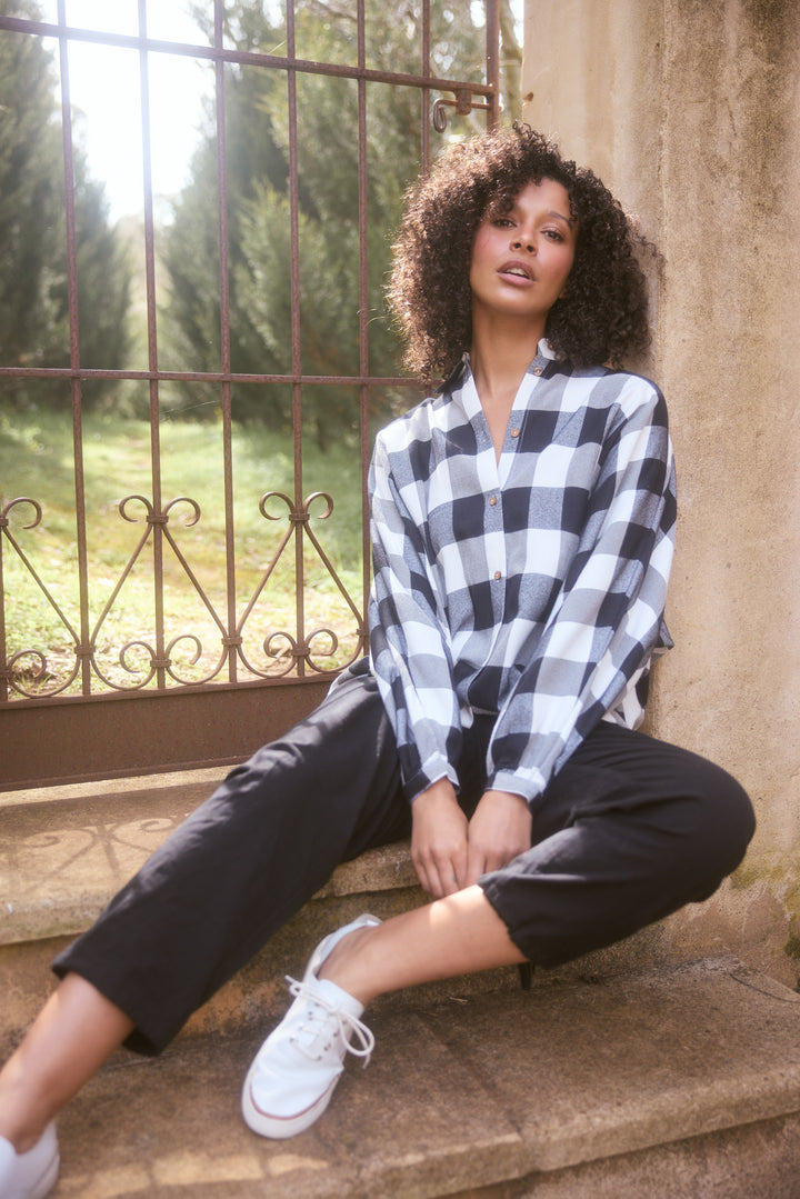 Woman sitting on steps wearing a checkered shirt and black pants, with a scenic background.