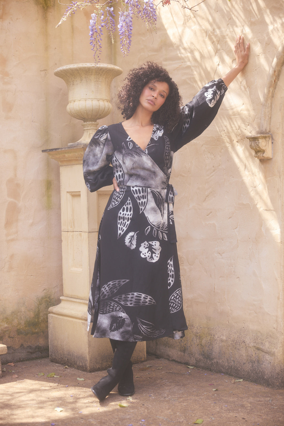 Woman in a black floral dress standing against a stone wall with wisteria.