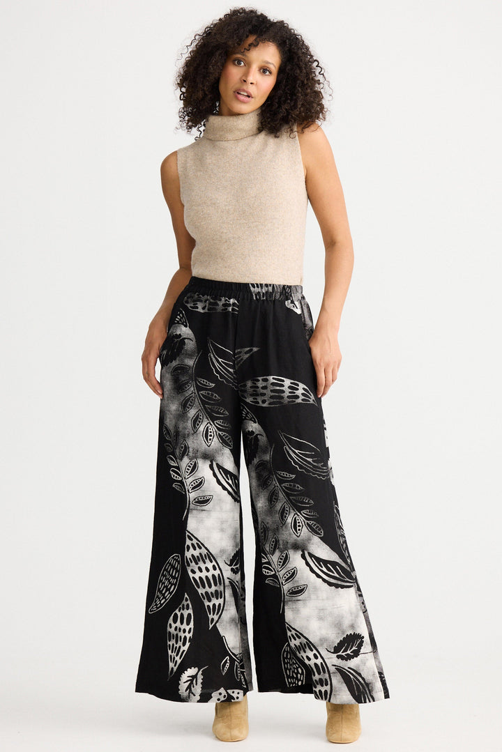 Woman wearing a sleeveless beige top and black patterned pants on a white background