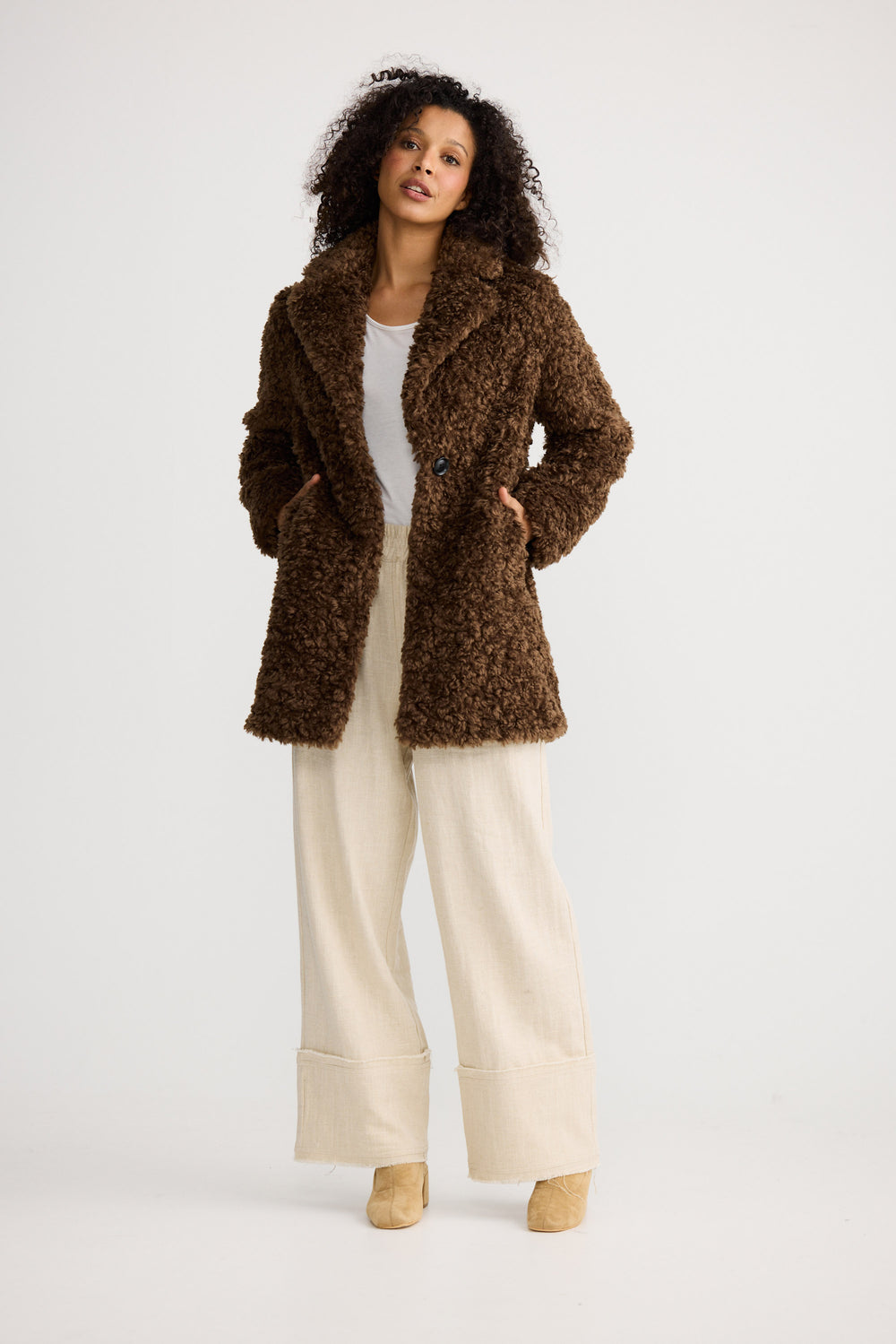 Woman wearing a brown textured coat with white pants on a light background