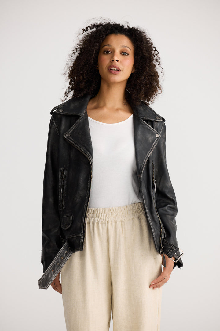 Woman wearing a black leather jacket, white top, and beige pants on a white background