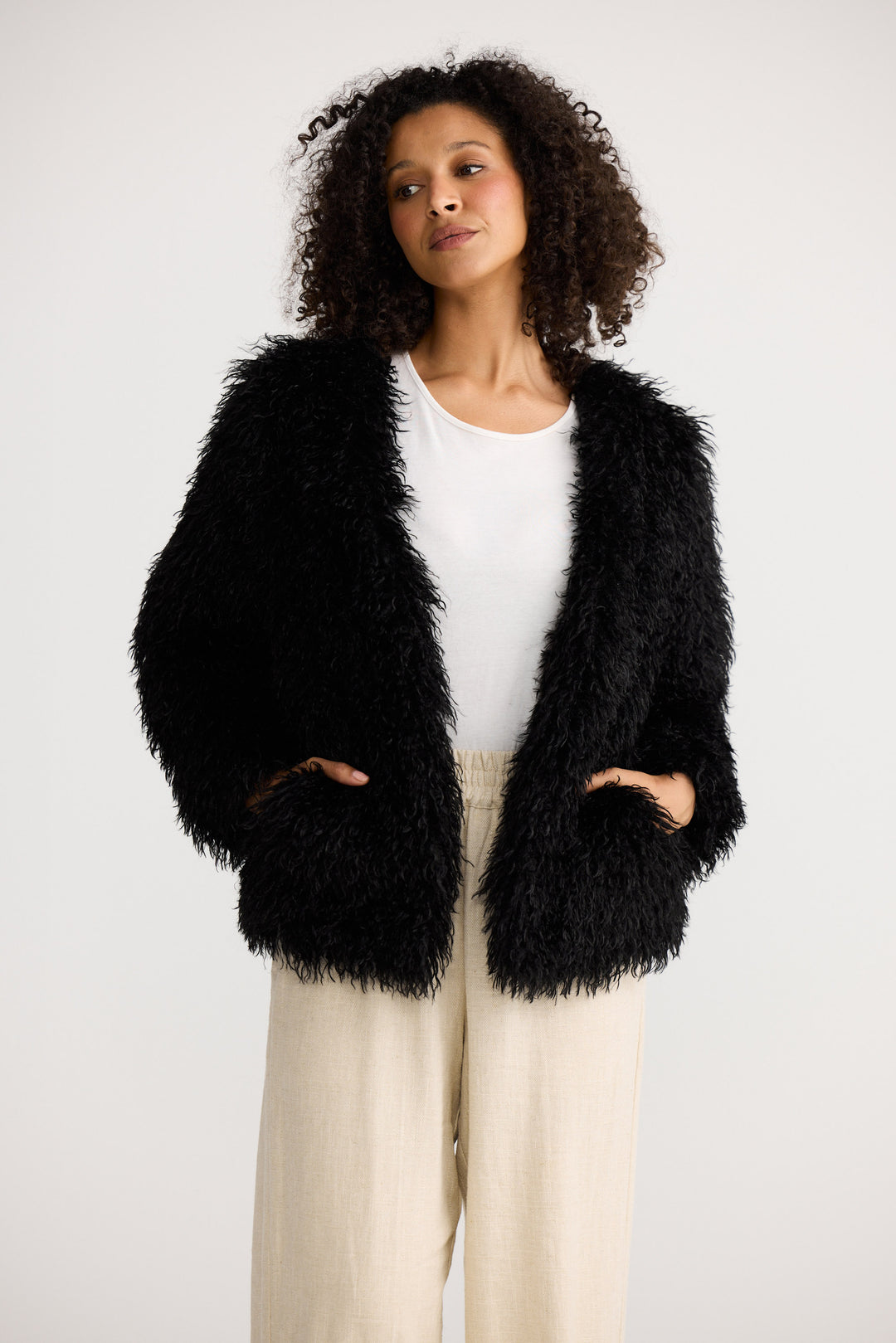 Woman wearing a black fluffy jacket over a white top and beige pants on a white background