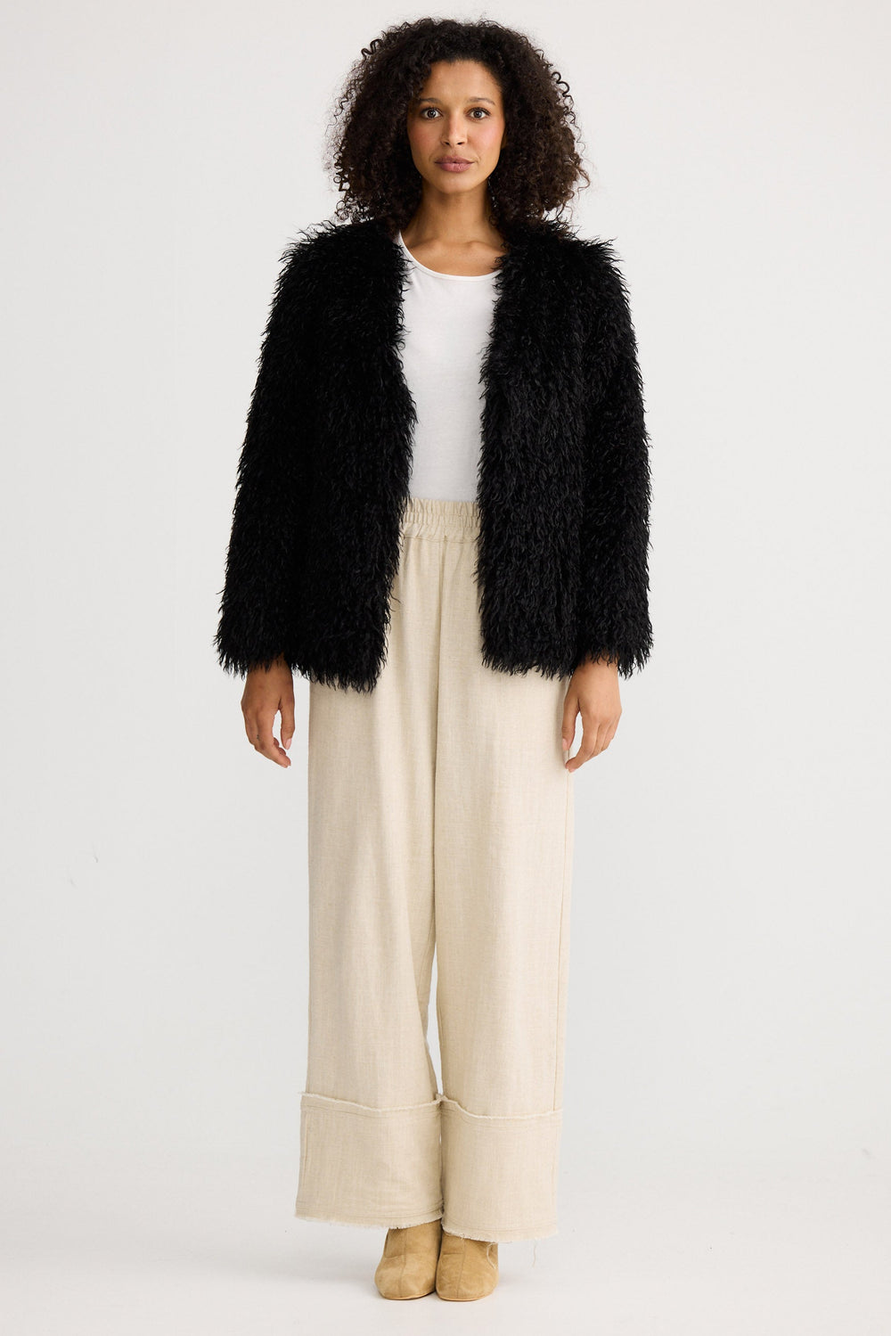 Woman wearing a black fluffy jacket, white top, and beige pants on a light gray background