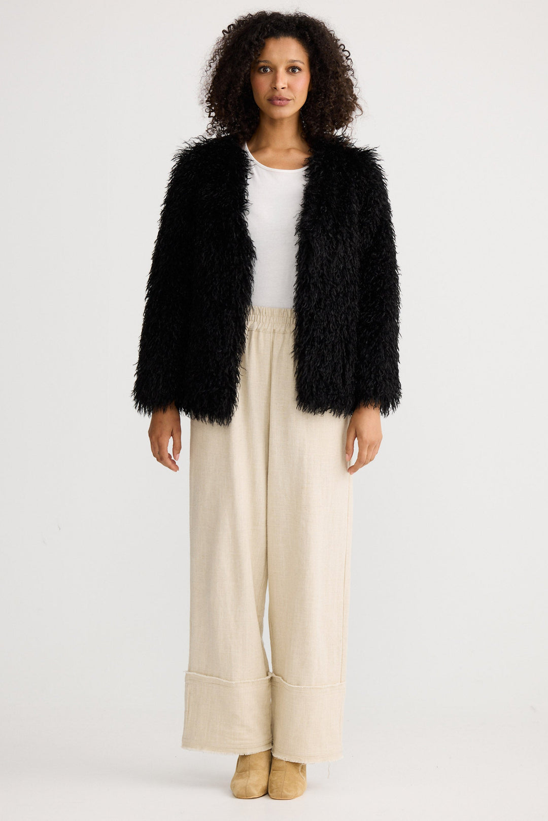 Woman wearing a black fluffy jacket, white top, and beige pants on a light gray background