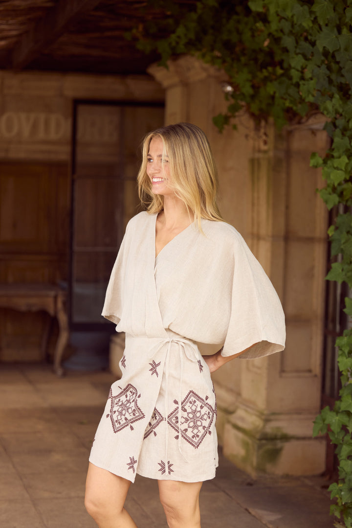 A woman wearing a cream-colored wrap robe and matching shorts with brown geometric patterns, standing in front of a stone building with green ivy.