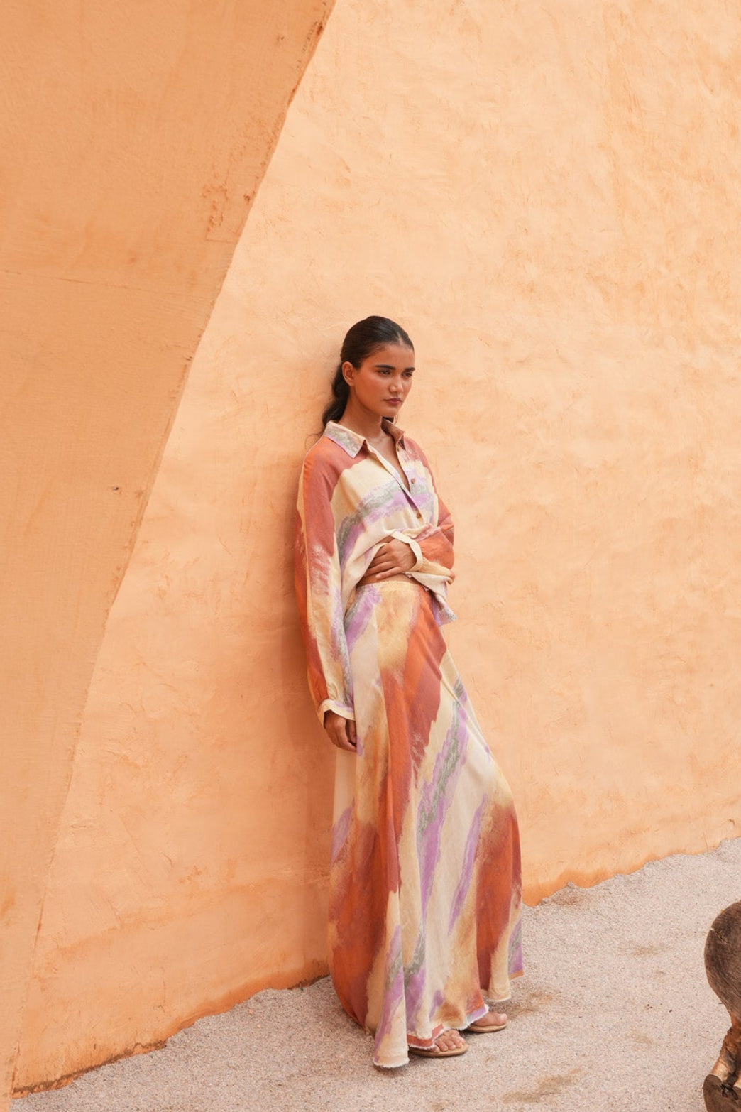 A woman wearing a long, flowing multicolor dress with orange, pink, and purple tones stands against a large, textured ochre-colored wall in an outdoor setting.