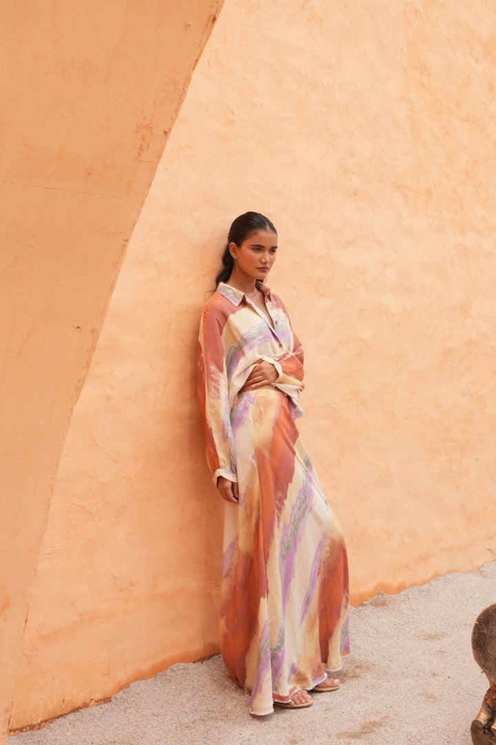 A woman wearing a long, flowing multicolor dress with orange, pink, and purple tones stands against a large, textured ochre-colored wall in an outdoor setting.