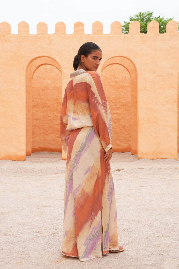 A woman stands outdoors in front of a peach-colored stucco wall with arches, wearing a long flowing dress with an abstract brushstroke pattern in orange, beige, cream, and lavender.