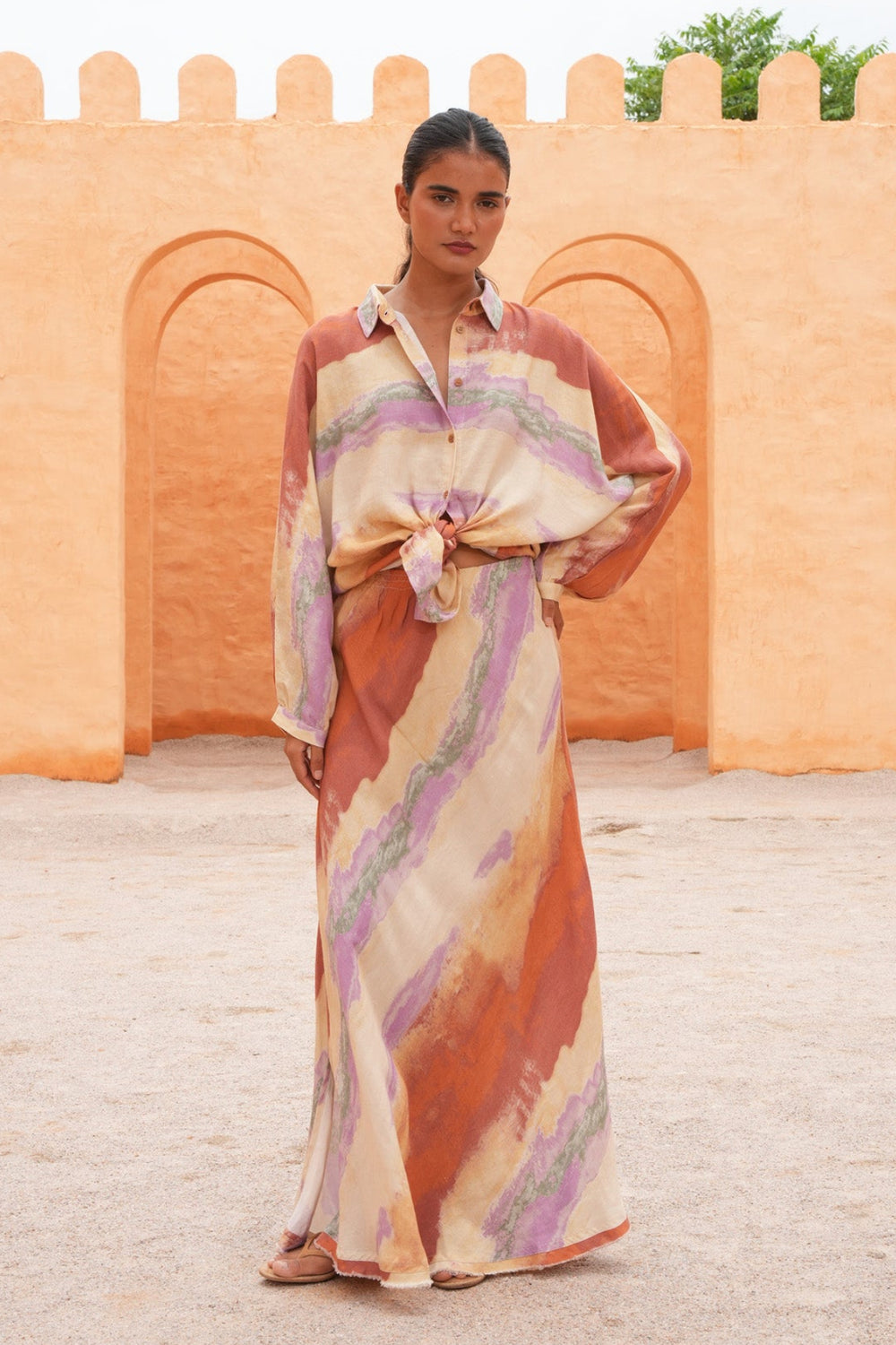 A woman stands outdoors in front of a warm orange adobe wall, wearing a long-sleeve collared maxi dress with a tie-front waist and an abstract brushstroke pattern in beige, rust orange, and lavender.