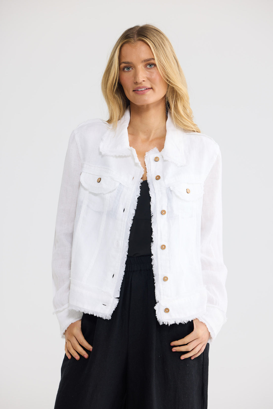 A blonde woman wearing a white frayed-edge denim jacket with gold-tone buttons over a black dress, standing against a light gray studio background.