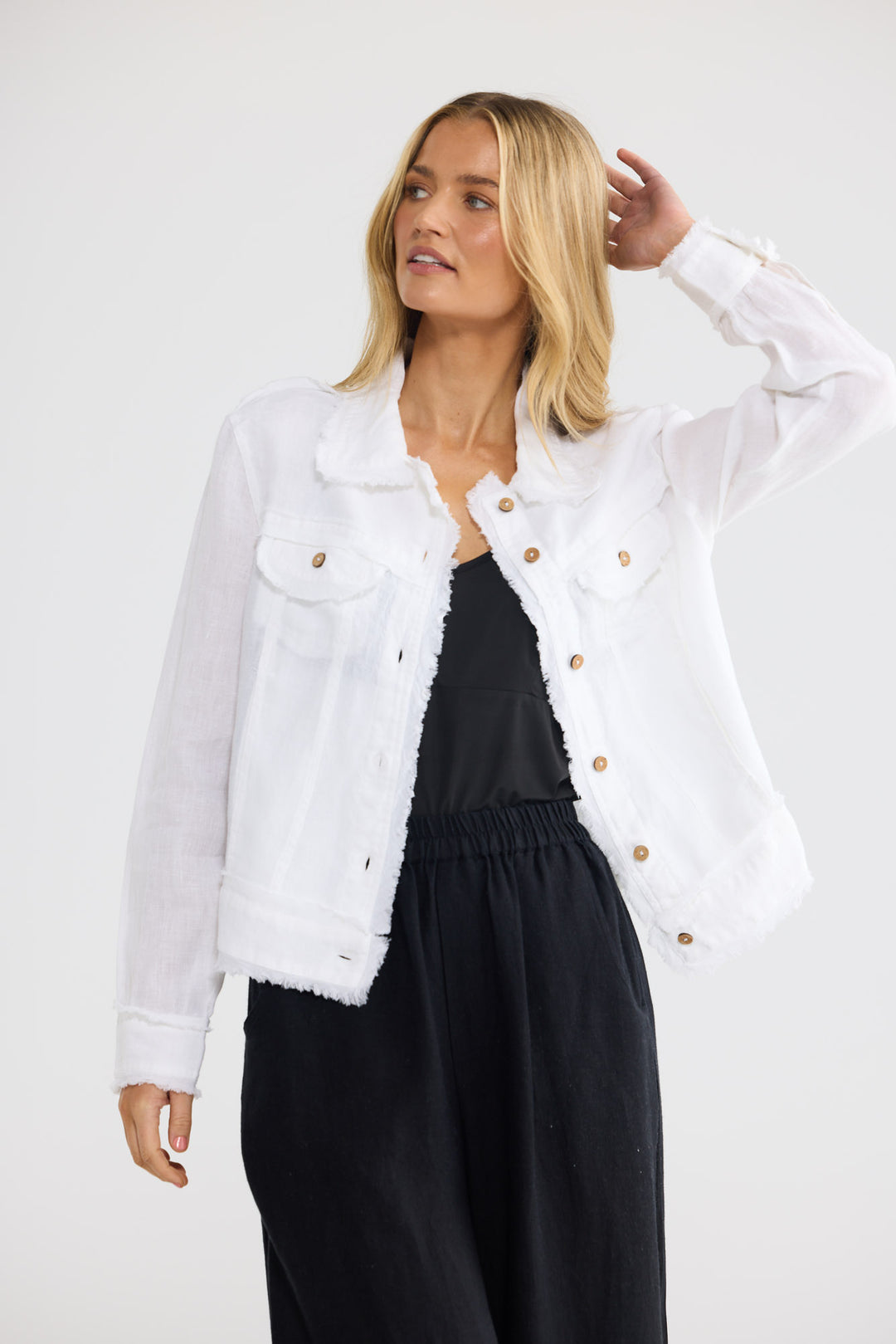 A blonde woman wearing a white frayed-edge denim jacket over a black top and black wide-leg pants, posing against a light gray studio background.