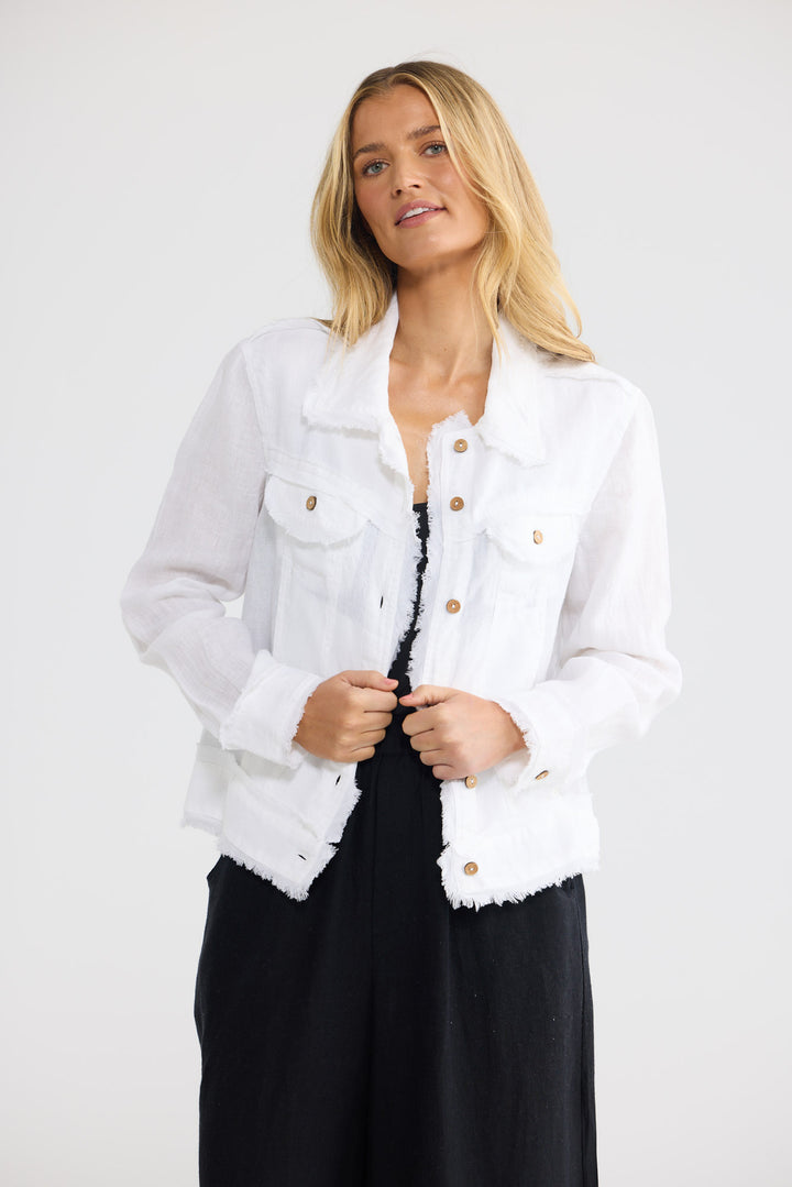 A blonde woman wearing a white frayed-edge denim jacket with gold-tone buttons, styled over a black top and black wide-leg pants, photographed against a light gray studio background.